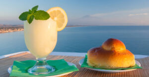 immagine che mostra un bicchiere di granita di limoni con acccanto una Brioche e come sfondo una foto del mare.